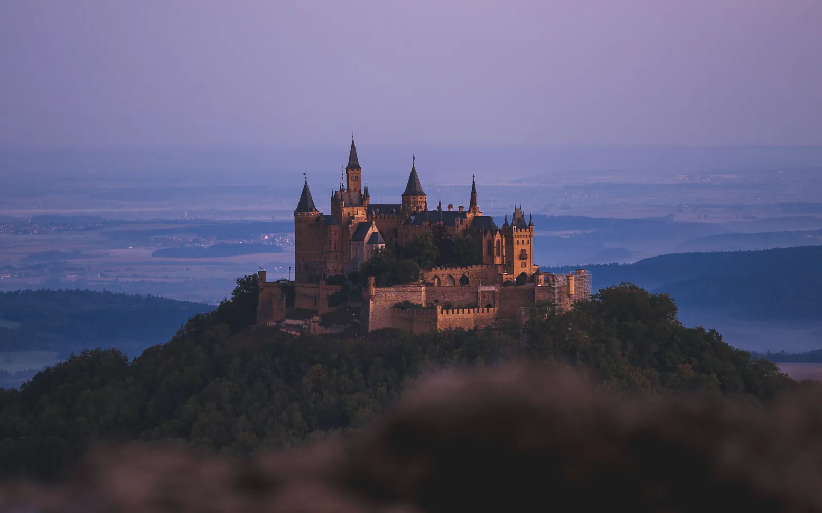 Burg Hohenzollern on its wooded hilltop at dusk, towers lit above the Swabian plain